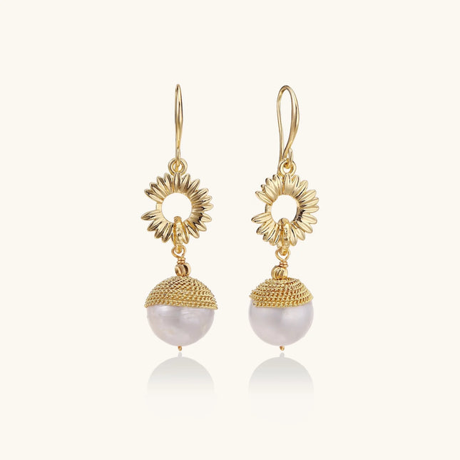 Gold sunburst charm earrings with baroque pearl drops on French hooks against cream background