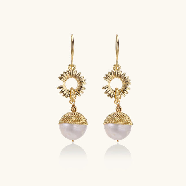 Gold sunburst charm earrings with baroque pearl drops on French hooks against cream background