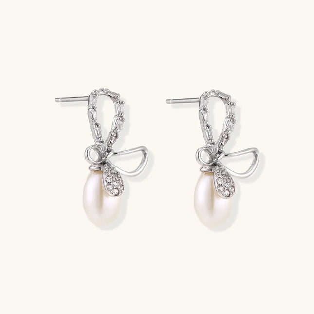 Silver bow-shaped stud earrings with baguette crystals and dangling white freshwater pearls on cream background
